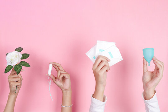 Four Hands Hold Various Menstrual Products And Flower Against Pink Background Displaying Feminine Hygiene Choices