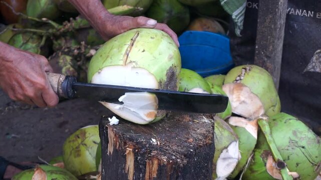 Hands of a vendor peeling a young green coconut using a sharp machete at a traditional market.	