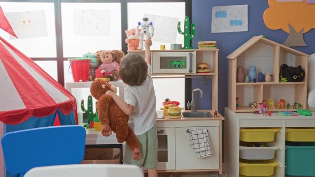 Boy holding plush monkey reaches up and points to a doll on a shelf in a preschool building playroom with toy kitchen, tent, and storage bins visible; curiosity imagination.