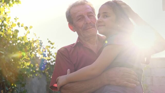 An elderly man embraces a young girl in a sunlit garden as they smile at each other, showing grandparent love, family warmth, trust, tenderness and a peaceful intergenerational bond.