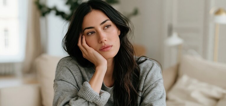 A thoughtful woman in a minimalist living room bathed in gentle natural light and subdued colors.