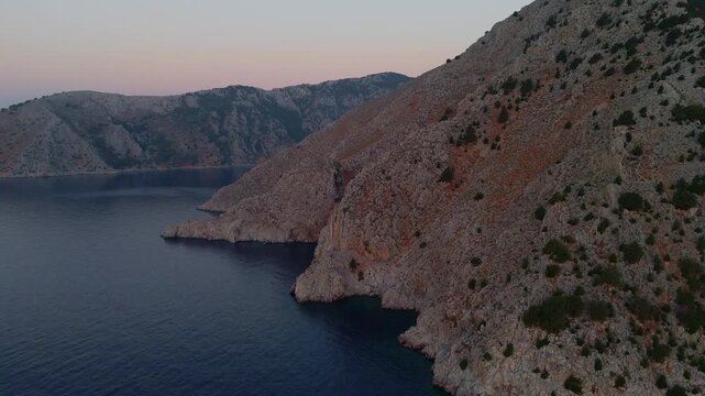 4K aerial shot of the dramatic rocky coastline in Symi Island, Greece during a calm golden hour sunrise.