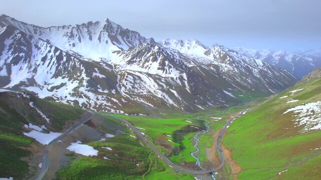 A vehicle driving along a winding mountain road flanked by towering snow-capped peaks; beautiful scenery in Xinjiang, China&mdash;the Duku Highway.