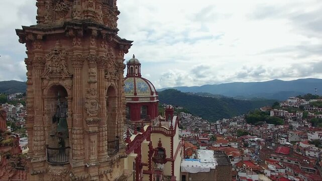 Aerial drone view rising and moving backward from Santa Prisca Church, revealing its baroque towers, tiled dome, and a panoramic vista of Taxco, Guerrero, Mexico.