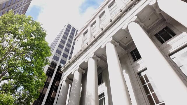 Majestic neoclassical columns of the Old Federal Reserve Bank building in San Francisco captured in a wide angle shot, with dappled sunlight, green trees, and modern glass offices towering nearby.