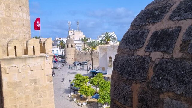 View from the fortress walls over the UNESCO-listed Medina of Sousse, Tunisia, where people cross the central square beneath a waving Tunisian flag on a sunny day
