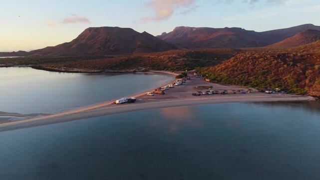 A cinematic wide-angle drone sequence flying low over calm, glassy waters to reveal the base of the Playa El Reques&oacute;n sandspit packed with beach campers under warm sunset light.
