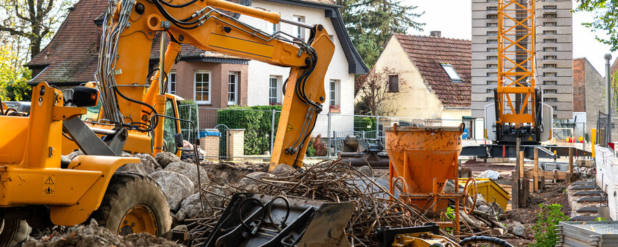 Yellow excavator works on urban construction site beside rubble, soil, concrete, fences, and residential houses. Hydraulic boom, crawler tracks, and debris show roadwork, renovation, infrastructure