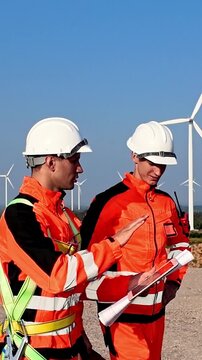 Two wind farm technicians in orange safety gear and hard hats review plans on site, with white turbines in the background under a clear blue sky.