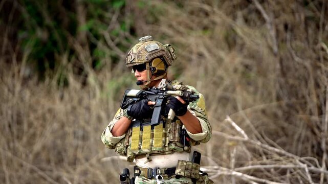 Soldier in camouflage and body armor aims a rifle during a field operation in dry brush, wearing helmet, headset, gloves, and tactical gear.