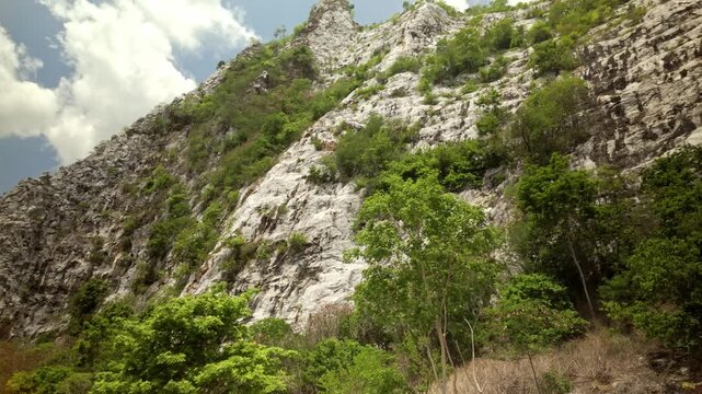 Rugged limestone cliff dotted with green trees and shrubs rises steeply under a bright, partly cloudy sky.