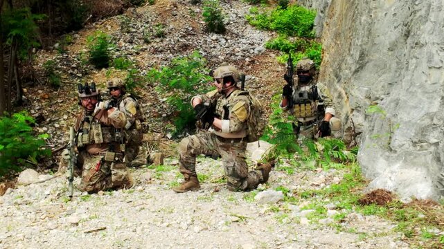 Armed soldiers in camouflage conduct a tactical patrol along a rocky hillside, coordinating positions and scanning the forested terrain during a field training exercise.