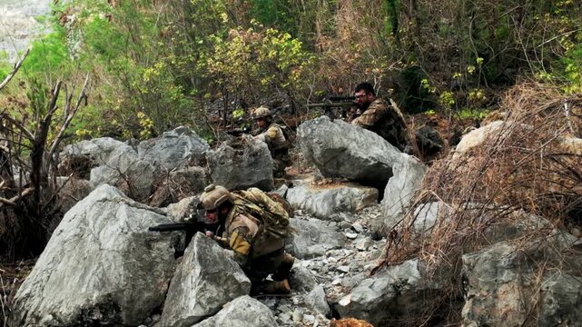 Camouflaged soldiers take cover among large rocks and sparse brush on a rugged hillside, aiming rifles during a tactical patrol in mountainous terrain.