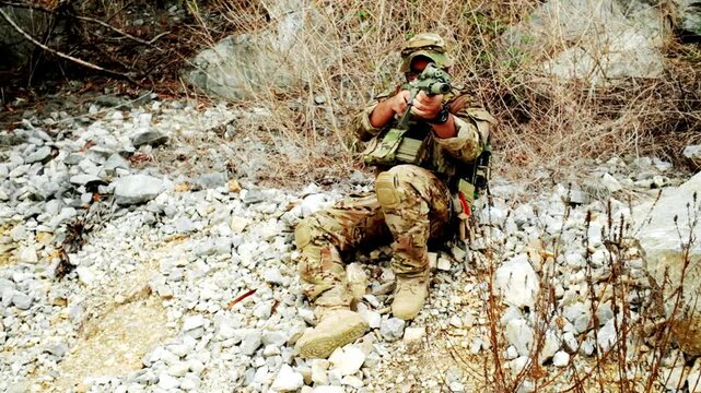Camouflaged soldier aims a rifle while seated among rocks and dry brush, blending into rugged terrain during a tactical operation.