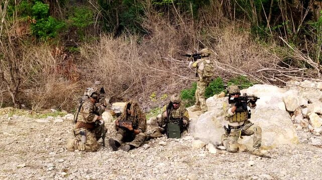 A tactical squad in camouflage conducts a field exercise on rocky terrain, with rifles ready and a radio operator coordinating amid sparse woodland.