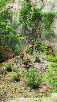 Camouflaged soldiers move in single file up a rocky ravine amid dense vegetation, conducting a tactical patrol or training exercise.