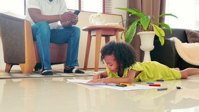 Smiling child in a green dress lies on a living room floor, drawing with colorful markers, while an adult sits nearby with a phone amid cozy furniture and houseplants.