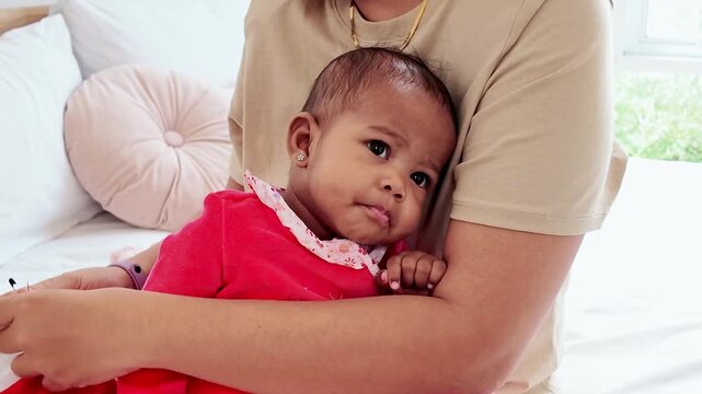 A baby in a red outfit rests in an adult"s arms at home, gazing at the camera on a cozy bed near a bright window, conveying warmth, safety, and tender bonding.