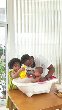 Smiling dad helps bathe a baby in a small tub on a table while an older child pours water, creating a warm, playful family moment in a bright, sunlit home.