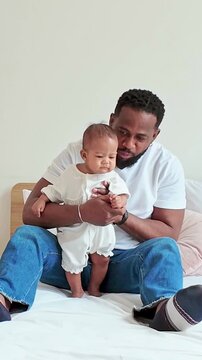 Smiling adult sitting on a bed helps a baby stand, sharing a tender moment in a bright, cozy bedroom.
