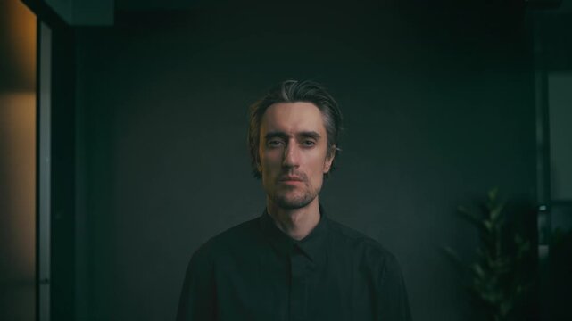 Serious young man with medium-length wavy hair in dark button-up shirt, indoor portrait with soft lighting, minimalist background, subtle plant, head shake no gesture