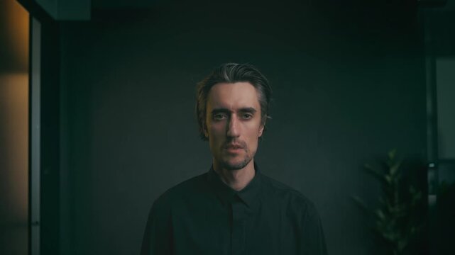 Moody indoor portrait of a man with medium-length hair in dark button-up shirt, standing against dim blurred background with subtle plant, minimalist professional look