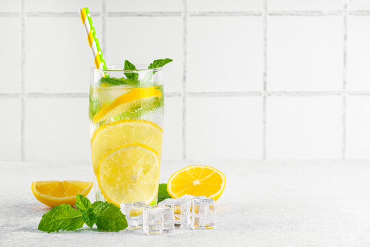 Refreshing homemade lemonade served in clear glass with fresh lemon slices, mint leaves, ice cubes and paper straws on a light kitchen counter against a white tile background with copy space.