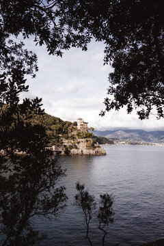 Elegant Mediterranean villa perched on a rocky coastline overlooking a calm bay, framed by lush tree branches under a dramatic cloudy sky. A peaceful coastal landscape with timeless Italian charm. Por
