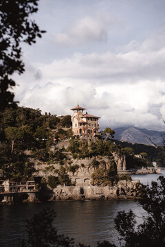 Elegant Mediterranean villa perched on rocky coastline overlooking calm bay, framed by lush tree branches under dramatic cloudy sky. peaceful coastal landscape timeless Italian charm Portofino