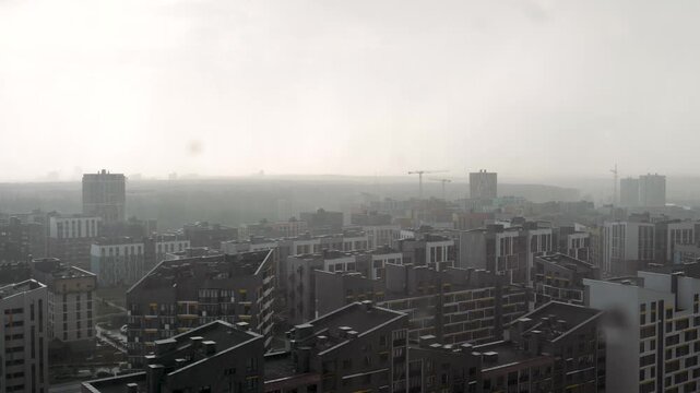 Dreary cityscape with modern apartment buildings and high rises under heavy rainfall on gloomy, overcast day. Raindrops are visible falling against the backdrop of urban landscape