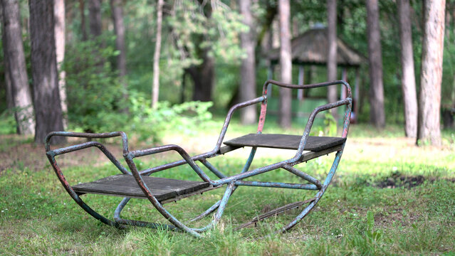 Old rusty swing gently sways in a peaceful forest clearing. Vintage abandoned playground bench rocking softly among trees on a sunny summer day.