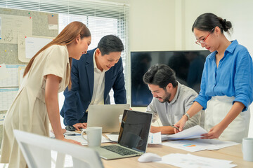 Fototapeta premium Diverse Asian business colleagues laughing during casual office discussion, global sales data map on screen, coffee mugs and documents on table, authentic candid corporate teamwork moment