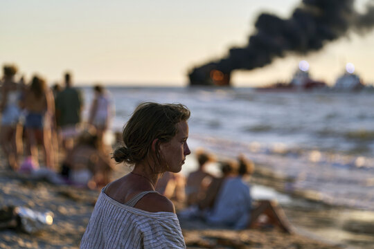 A contemplative woman gazes at the sea, oblivious to the boat fire in the distance, symbolizing the innocence of leisure threatened by unforeseen events.