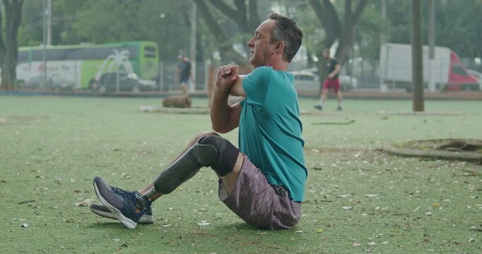 Man with a lower limb prosthesis performing a seated triceps stretch on grass in a public park, promoting adaptive exercise, physical rehabilitation, upper body mobility, flexibility, resilience,