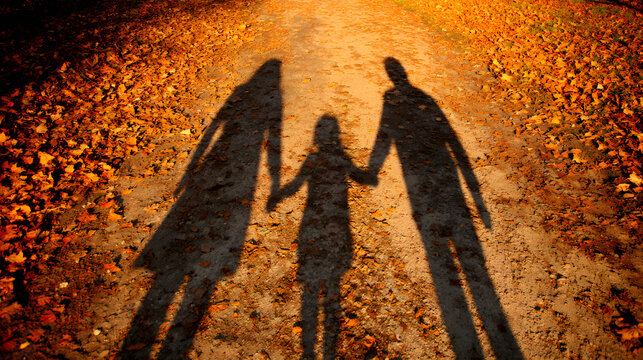Long shadows of a family of three holding hands while walking along a gravel path covered in vibrant orange and yellow autumn leaves in the park