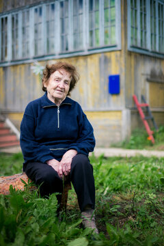 Environmental portrait of an elderly woman sitting on a tree log in front of a weathered wooden farmhouse, surrounded by green grass in a quiet rural setting.