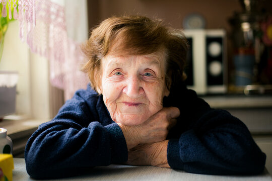Close portrait of an elderly woman resting her folded arms on a kitchen table, illuminated by soft window light in a quiet home interior.