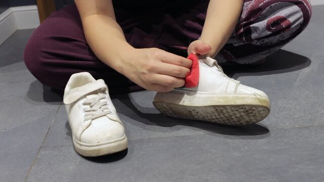 A white sneaker is cleaned with a red sponge on an indoor tiled floor with water nearby.