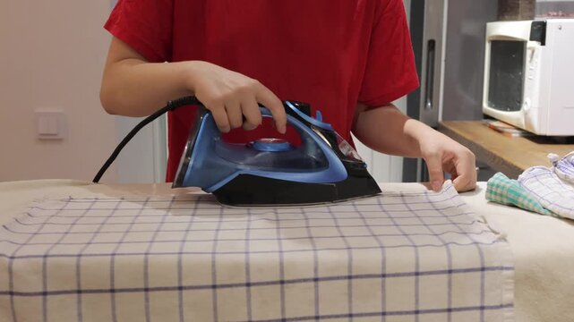 A steam iron moves over a checked kitchen towel while a person steadies the fabric on an ironing board.