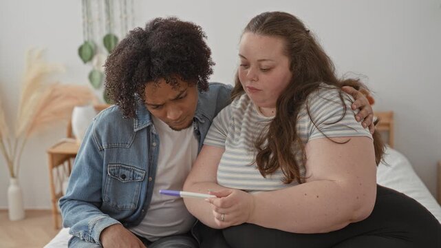 Woman holding pregnancy test on bed, man sitting beside her with arm around shoulder in a building; support concern.