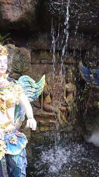 Vertical video of winged kinnara statue beside a waterfall and carved relief at the Blue Temple