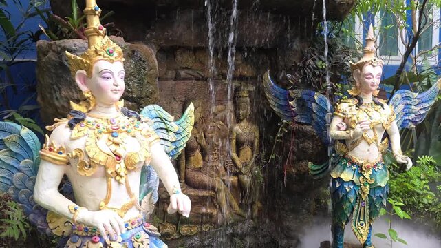 Slow motion vlog footage of winged kinnara statues beside a waterfall behind the Blue Temple
