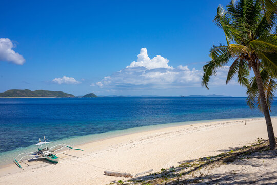 Pristine white sand beach with a palm tree and traditional outrigger boat by the shore and islands on the horizon.
