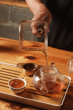 Hand pouring hot amber tea from a glass pitcher into a glass teapot on a wooden gongfu tea tray during a traditional tea ceremony.