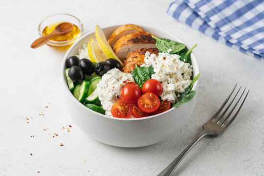 Healthy high protein Mediterranean chicken bowl with rice, cucumber, cherry tomatoes, feta cheese, olives, spinach and creamy yogurt sauce on light background with copy space