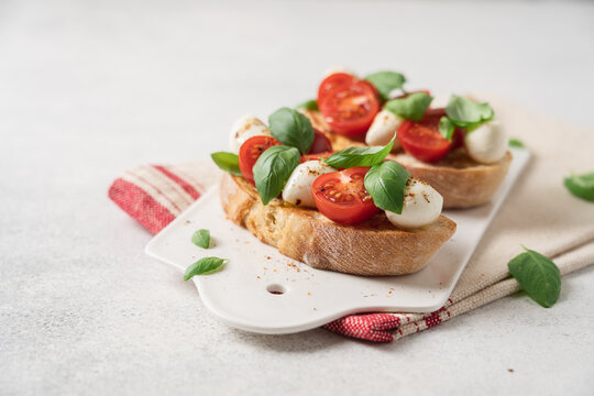 Bruschetta Caprese with cherry tomatoes mozzarella cheese and fresh basil on white ceramic board over light background, traditional Italian cuisine appetizer concept