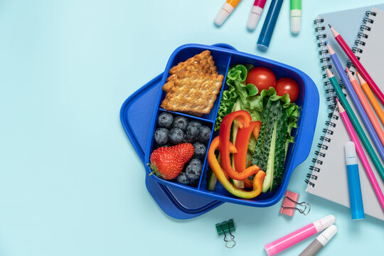 Blue school lunch box with berries crackers and vegetables surrounded by stationery on blue background, top view back to school concept