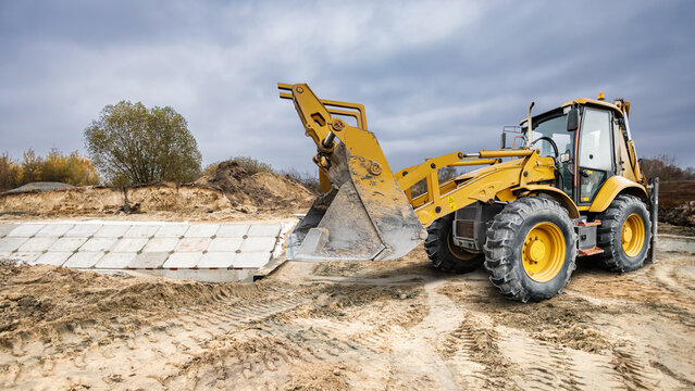 A universal excavator loader operates on a construction site. It moves materials and digs into sandy soil under a cloudy sky at an outdoor location