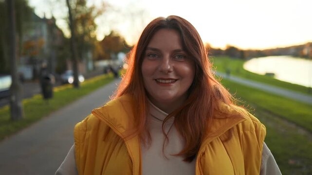 A woman wearing a yellow jacket stands and smiles on a pathway surrounded by grass and trees, captured from a slightly elevated frontal view. This commercial stock footage is usec.