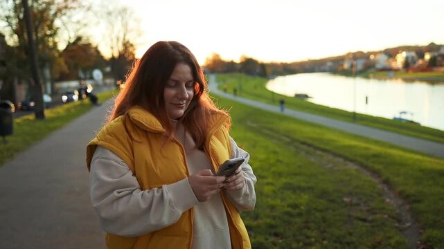 A woman in a yellow jacket is walking and smiling in a park by a lake, surrounded by trees and grassy areas. The scene is captured from a side view at a slightly low angle, emphasizing her.
